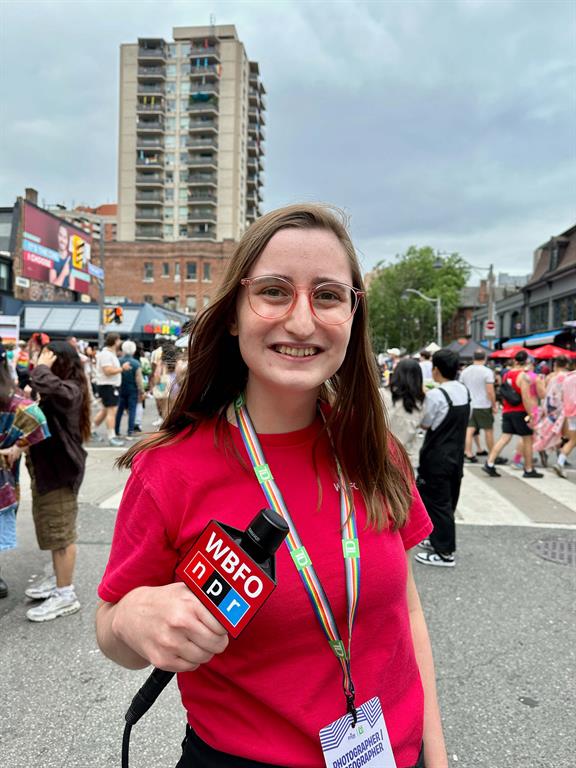 Emyle Watkins is posing for a photo with a reporter mic at Pride Toronto." longdesc="Emyle is smiling with her mic that reads WBFO, NPR. She is wearing a bright pink t-shirt with a pride-themed lanyard. The photo is taken on the streets of Toronto with a crowd and buildings in the background.